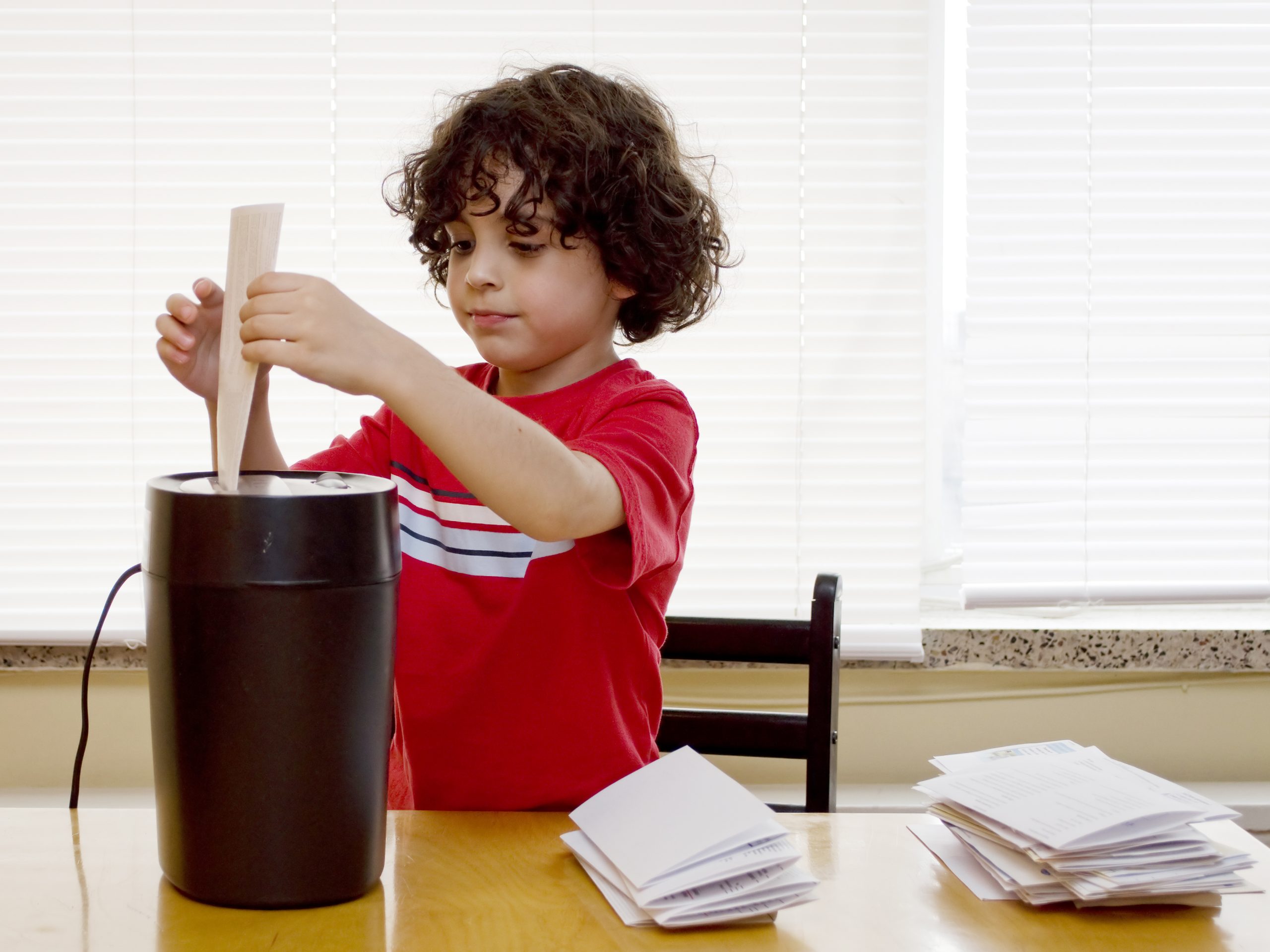 Little boy in red t shirt with dark curly loading a document into a paper shredder. 