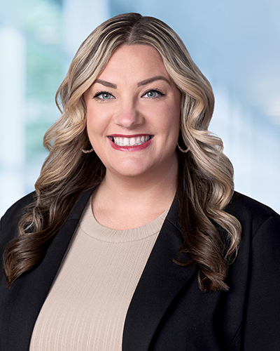 Woman with long blonde hair slightly curled with a tan shirt and black blazer on in front of a pale blue and green background.